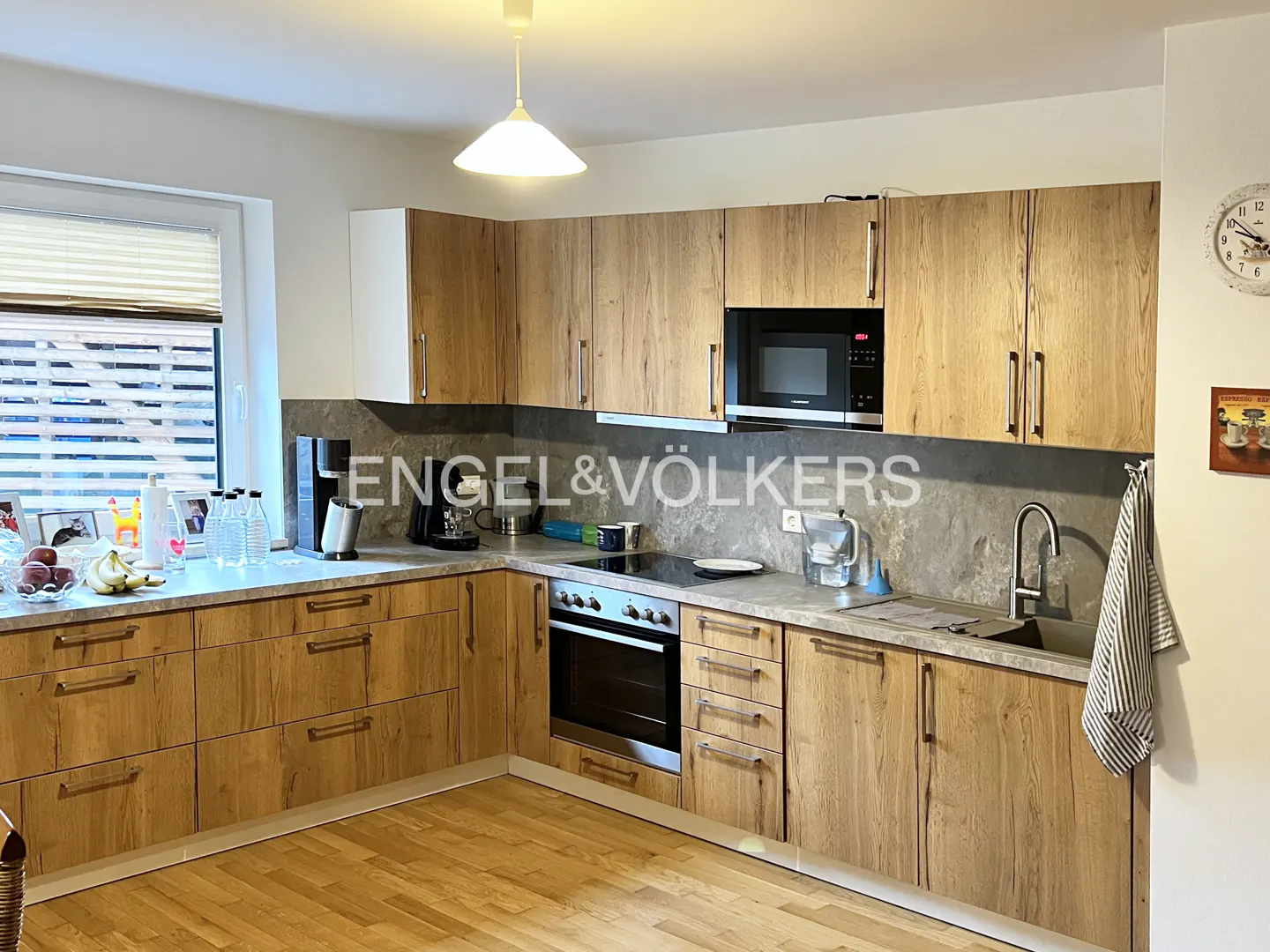 A kitchen with wood cabinets, gray countertops, and stainless steel appliances. A window is on the left, and a light fixture hangs from the ceiling.