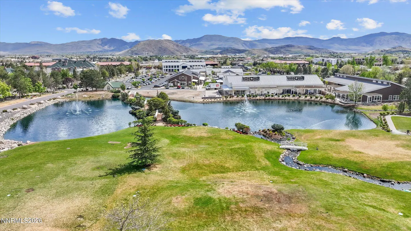 Aerial view of a pond with fountains, surrounded by green grass, buildings, and mountains in the background.