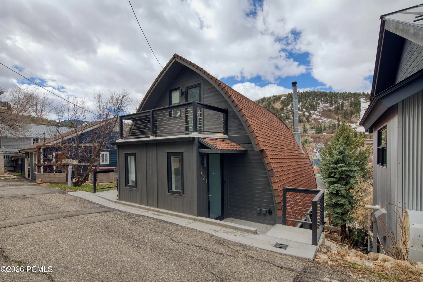 Exterior view of a modern gray house with a curved roof and a balcony, set on a hillside street.