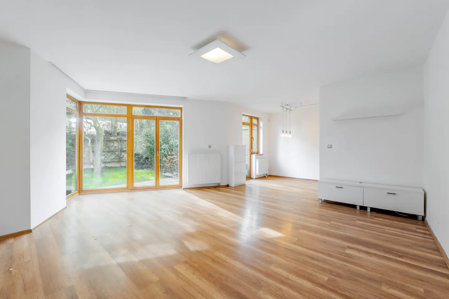 Bright, empty room with wood floors, white walls, and large windows overlooking a green yard. A white cabinet sits against the far wall.