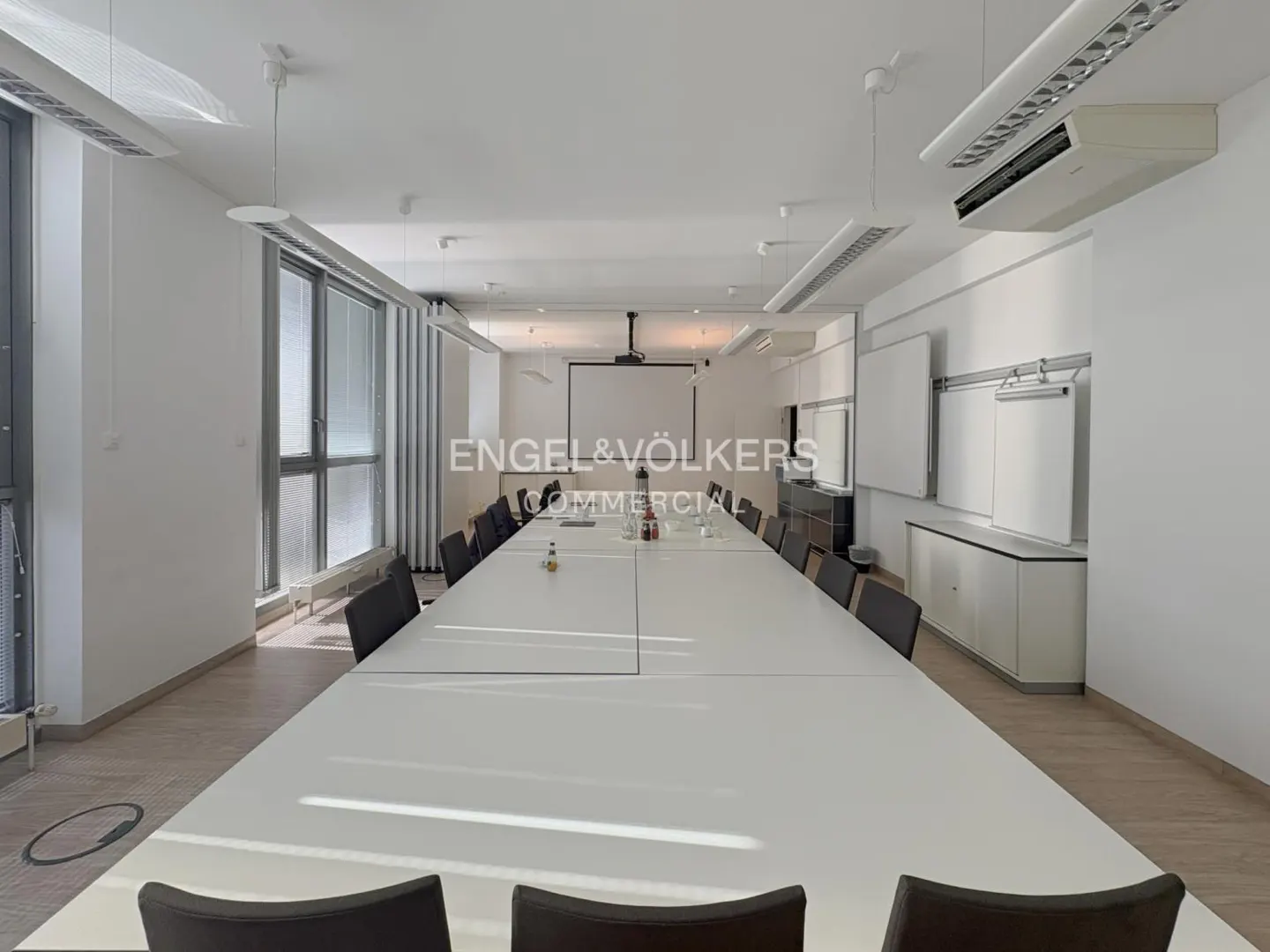 Bright, modern conference room with a long white table, dark chairs, projector screen, and whiteboards.
