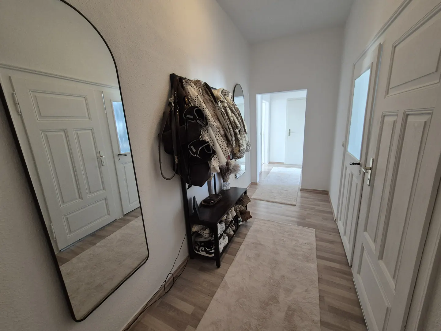Hallway with a full-length mirror, coat rack with jackets, and a shoe rack. The walls and doors are white, and there is a light-colored rug on the floor.