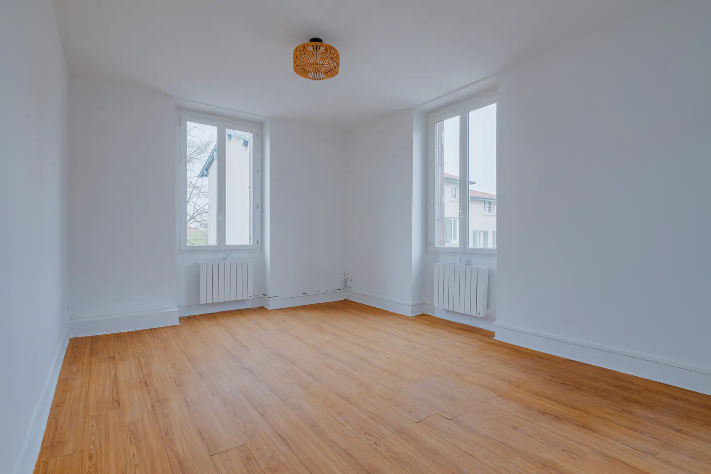Bright, empty room with wood floors, white walls, and two windows. A wicker light fixture hangs from the ceiling. Radiators are under each window.