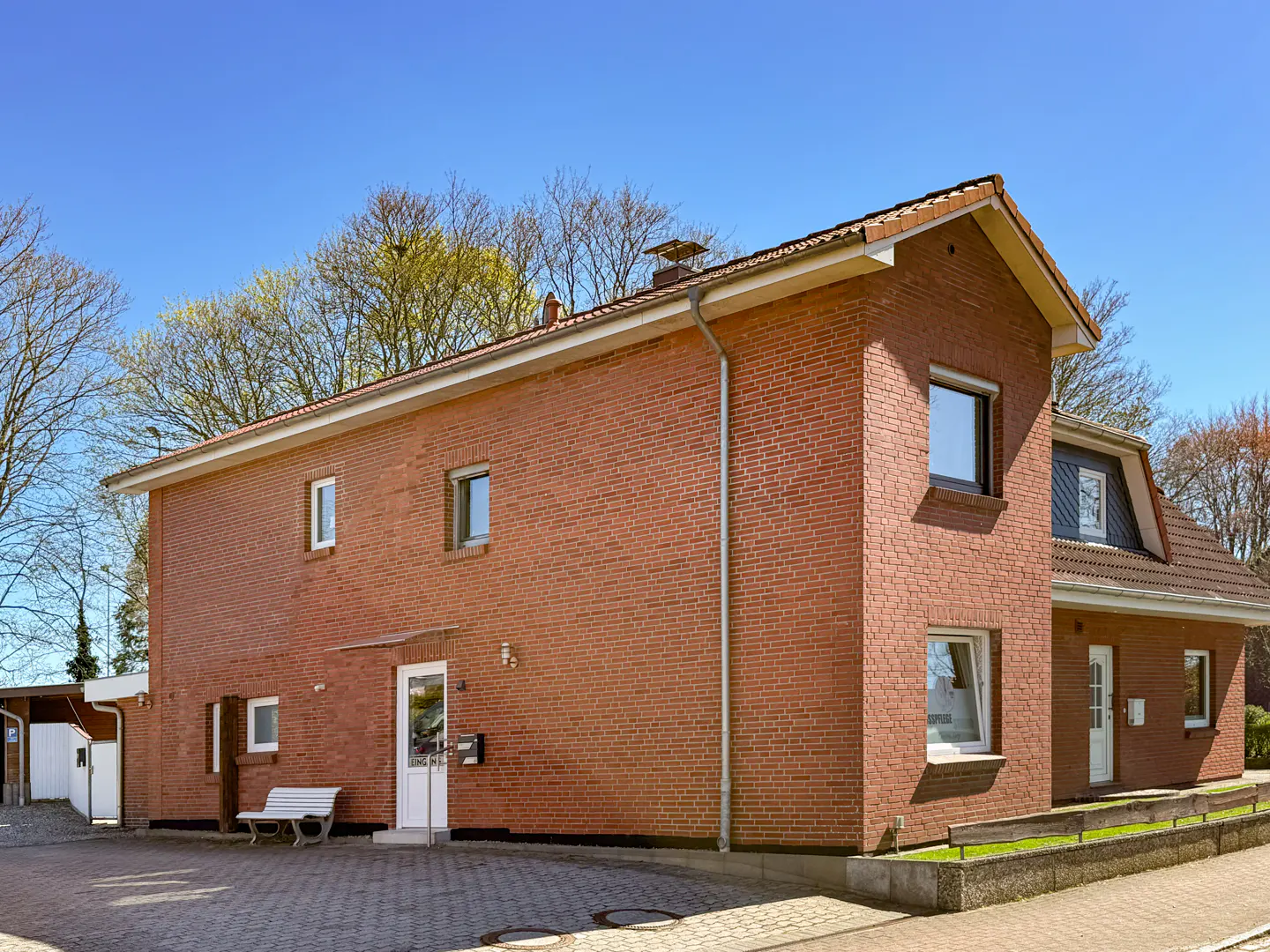 Two-story red brick house with white trim and a red tile roof under a clear blue sky. A white bench sits near the front door.