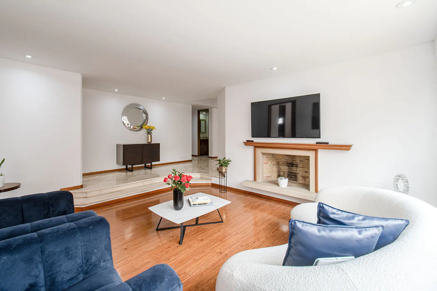 Bright living room with wood floors, white walls, blue sofa, and white curved sofa. A TV hangs above a fireplace. A marble landing leads to a hallway.