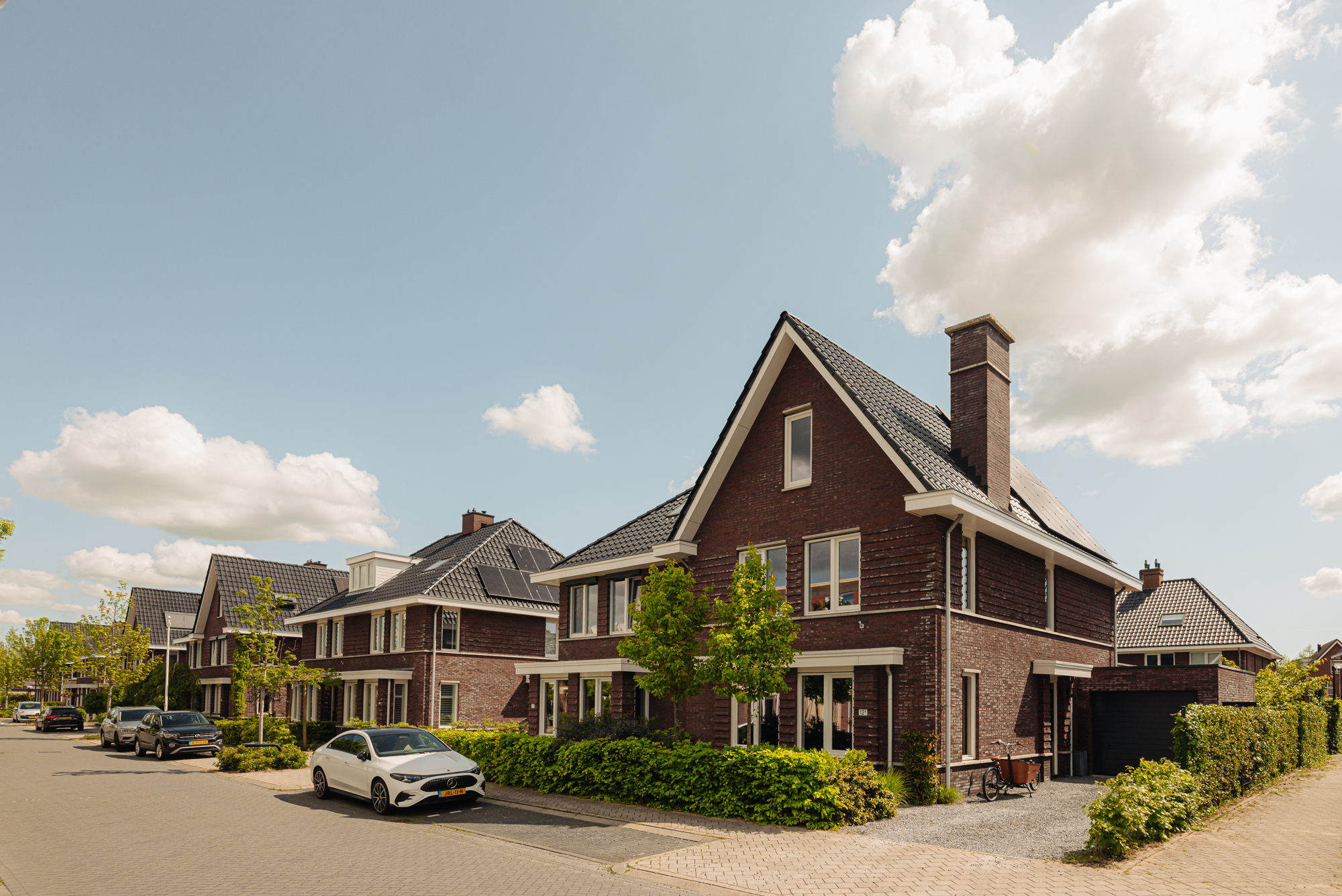 Row of brick houses with black roofs and solar panels under a blue sky with white clouds. Cars parked on the street.
