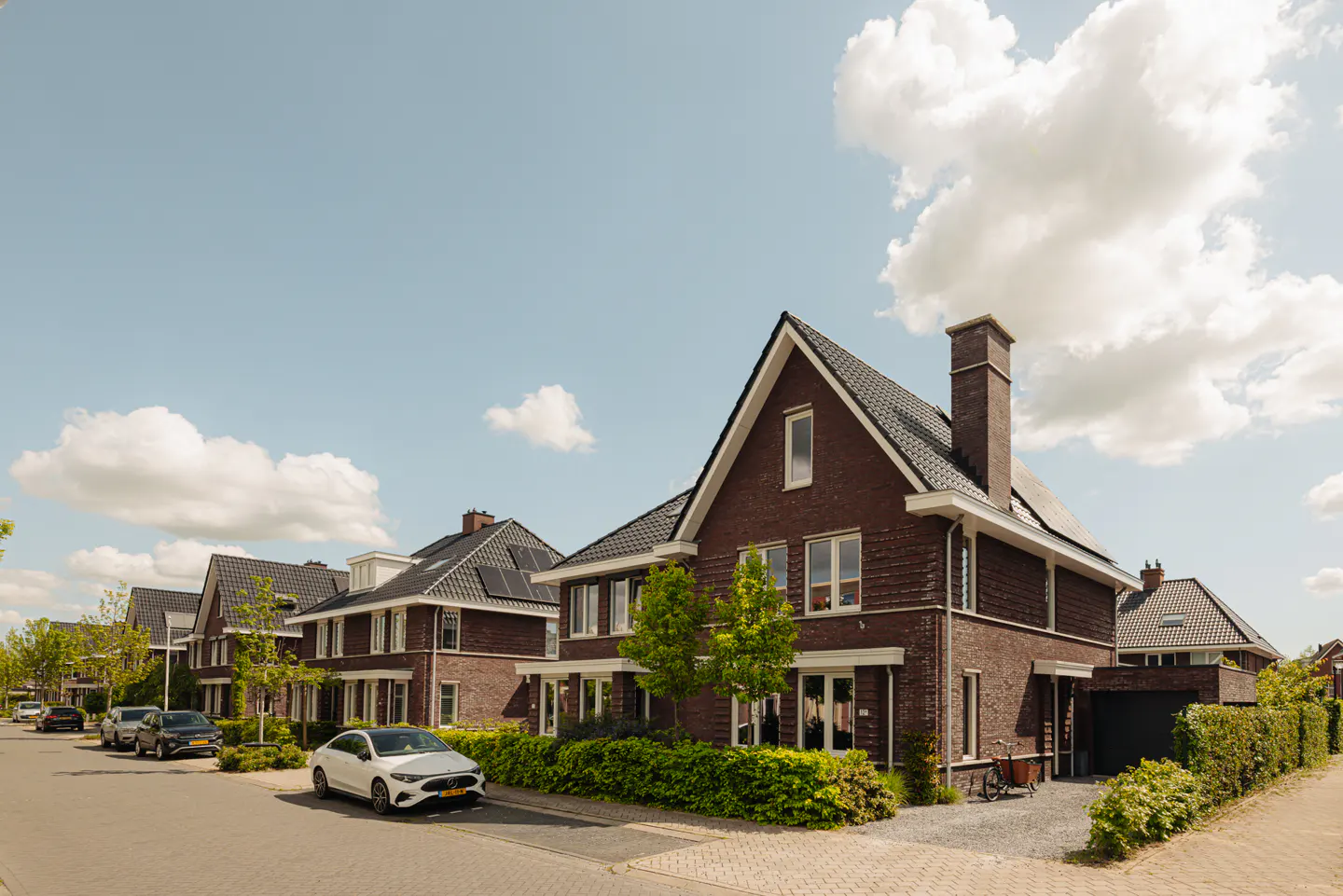 Row of brick houses with black roofs and solar panels under a blue sky with white clouds. Cars parked on the street.