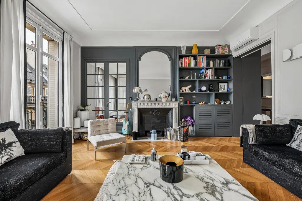 Living room with herringbone wood floors, black sofas, marble coffee table, fireplace, and built-in gray bookcase.