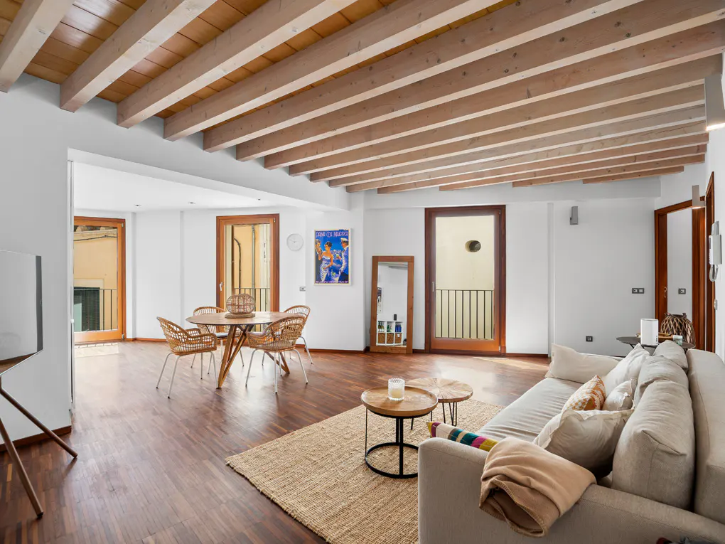Bright living room with wood floors, beamed ceiling, and white walls. A beige sofa sits on a jute rug, with a round table and wicker chairs in the background.