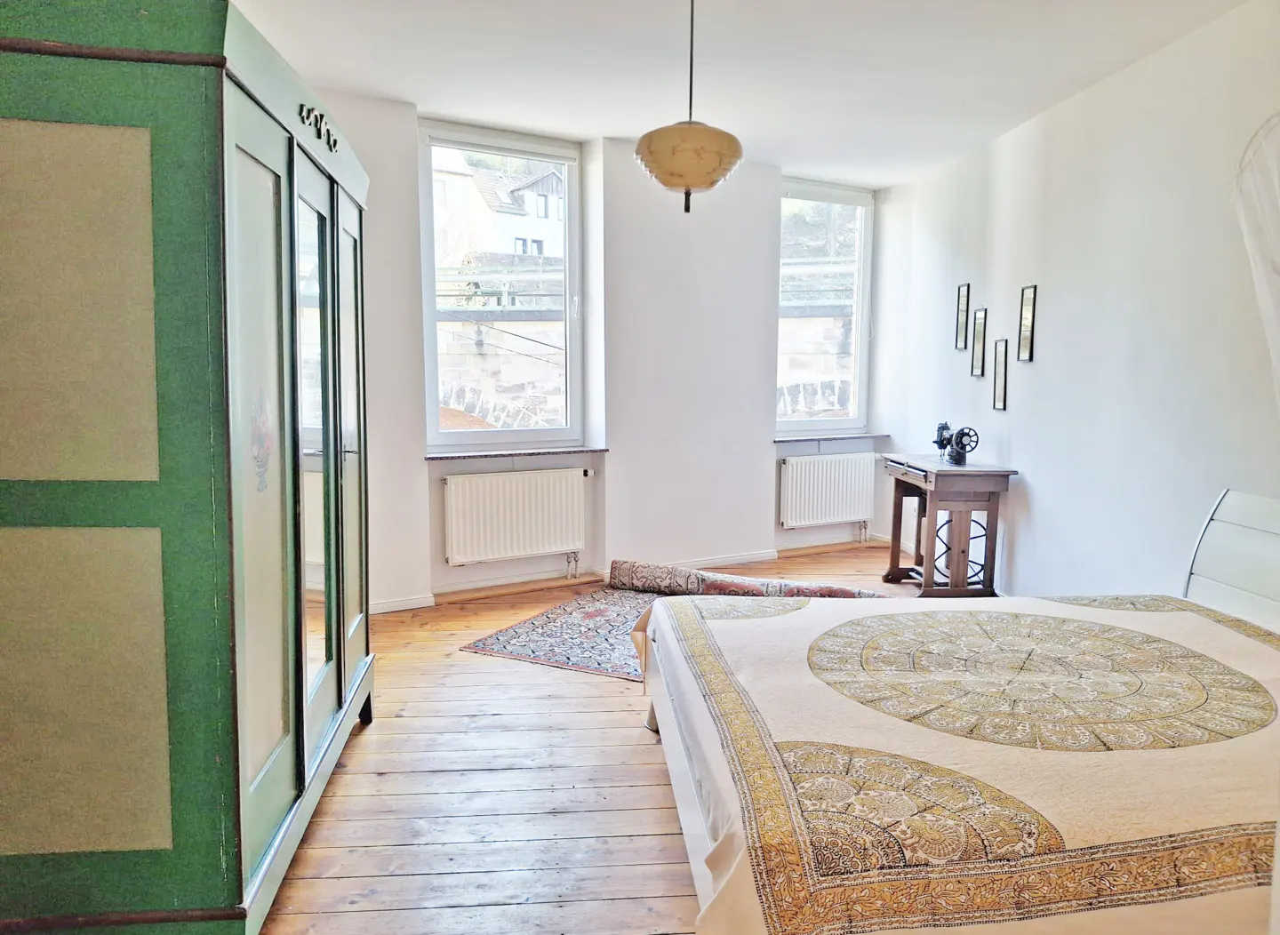 Bright bedroom with wood floors, white walls, and two windows. A green wardrobe stands to the left, and a bed with a patterned cover is in the foreground.