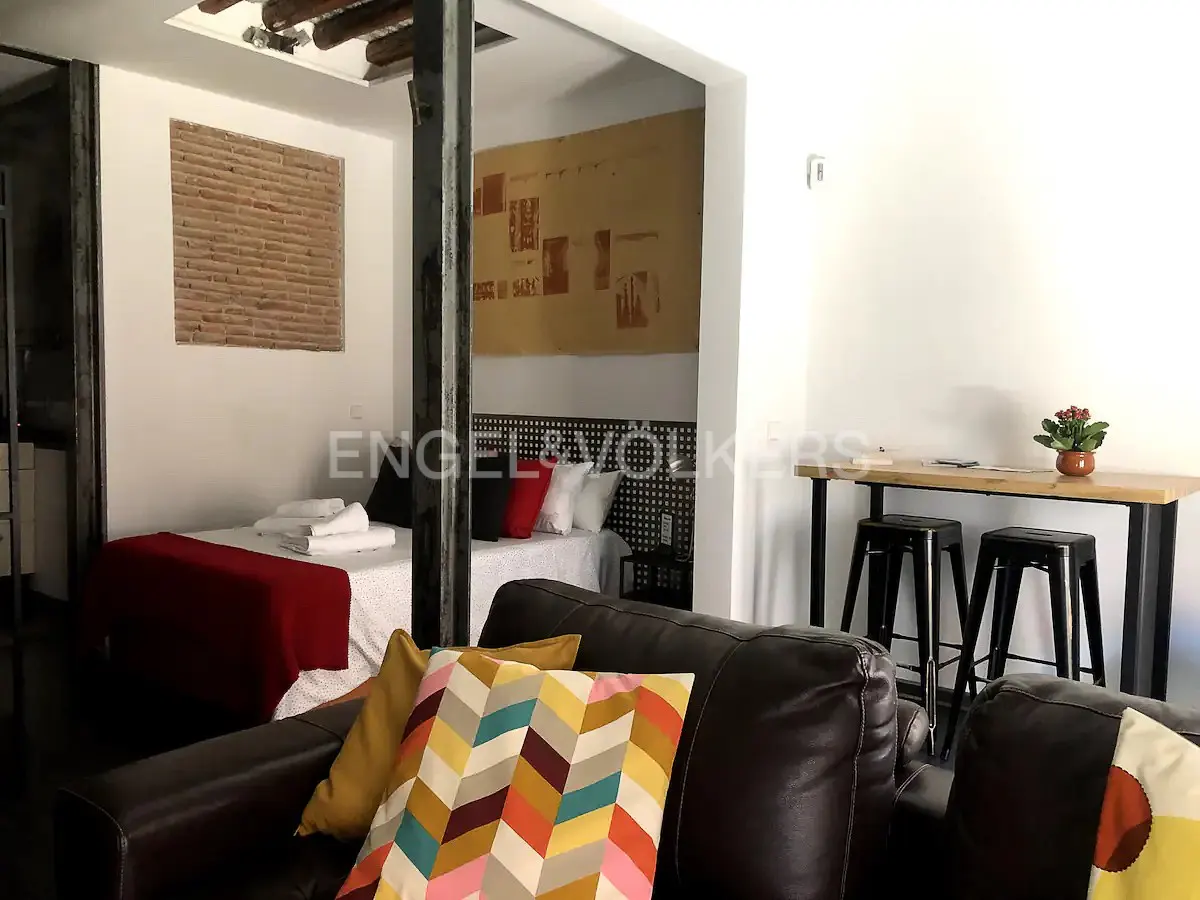 Interior view of a modern apartment with a bed, brown leather sofa with colorful pillows, and a high table with stools.
