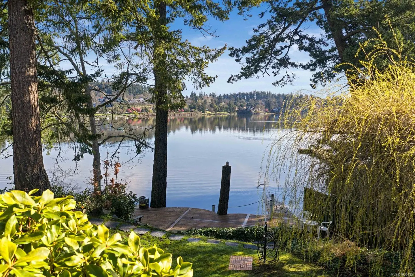 Waterfront property view with a wooden dock, trees, and a calm lake reflecting the sky. A small seating area is visible on the right.