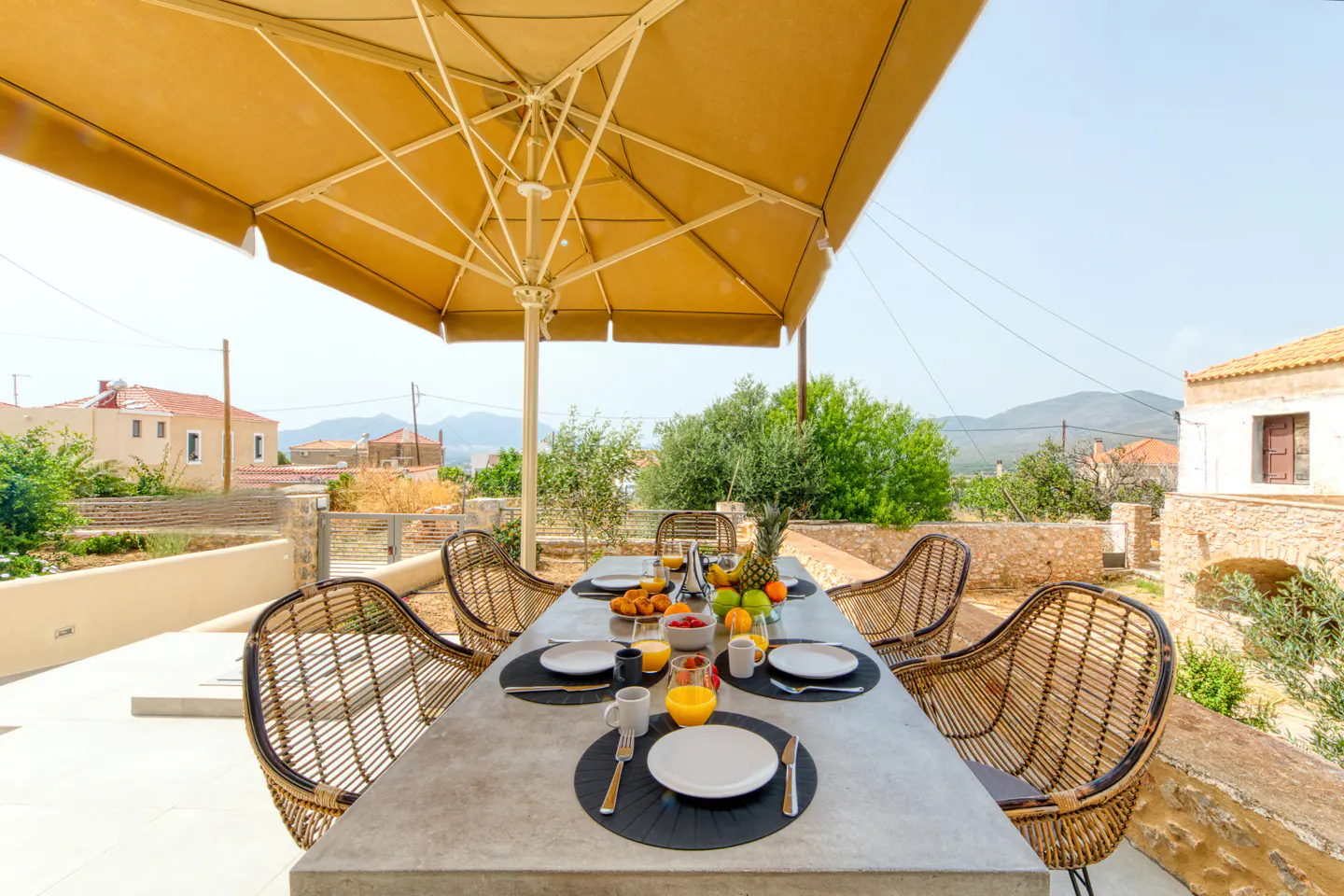 Outdoor dining area with a concrete table, wicker chairs, and a large tan umbrella. Breakfast is set on the table with fruit and juice.