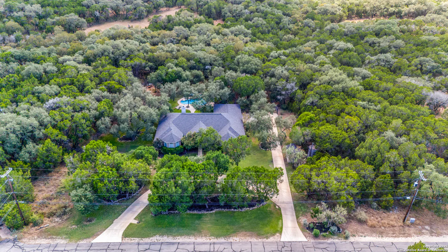 Aerial view of a house with a gray roof and a blue pool, surrounded by lush green trees. Two driveways lead to the house.