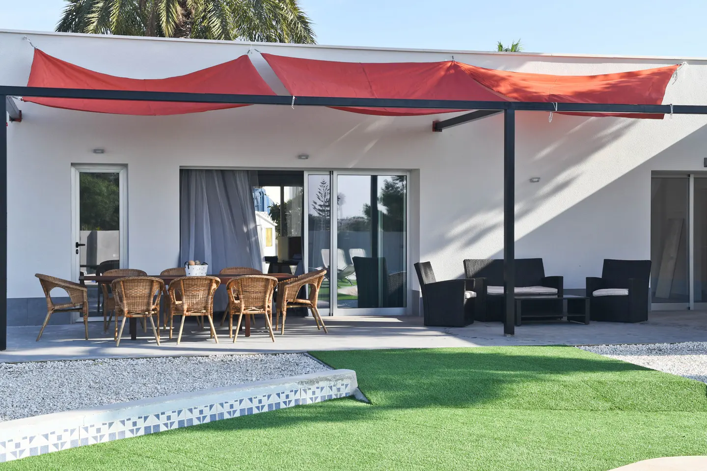 Outdoor patio with wicker dining set, black lounge furniture, and red sunshade. Green lawn and white building in background.
