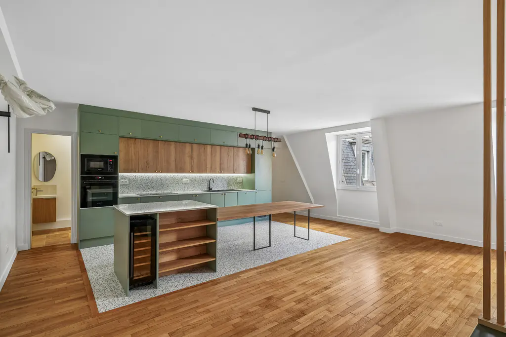 Bright, modern kitchen with green and wood cabinets, island with wine fridge, and long table on a speckled rug. Hardwood floors and white walls.