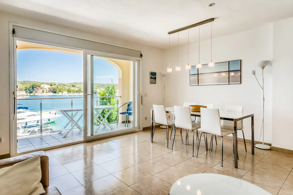 Bright dining room with a white table, chairs, and a balcony overlooking a blue ocean with boats.