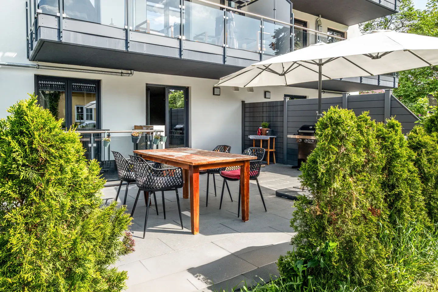 A patio with a wooden dining table, black chairs, a white umbrella, and a grill, framed by green hedges.