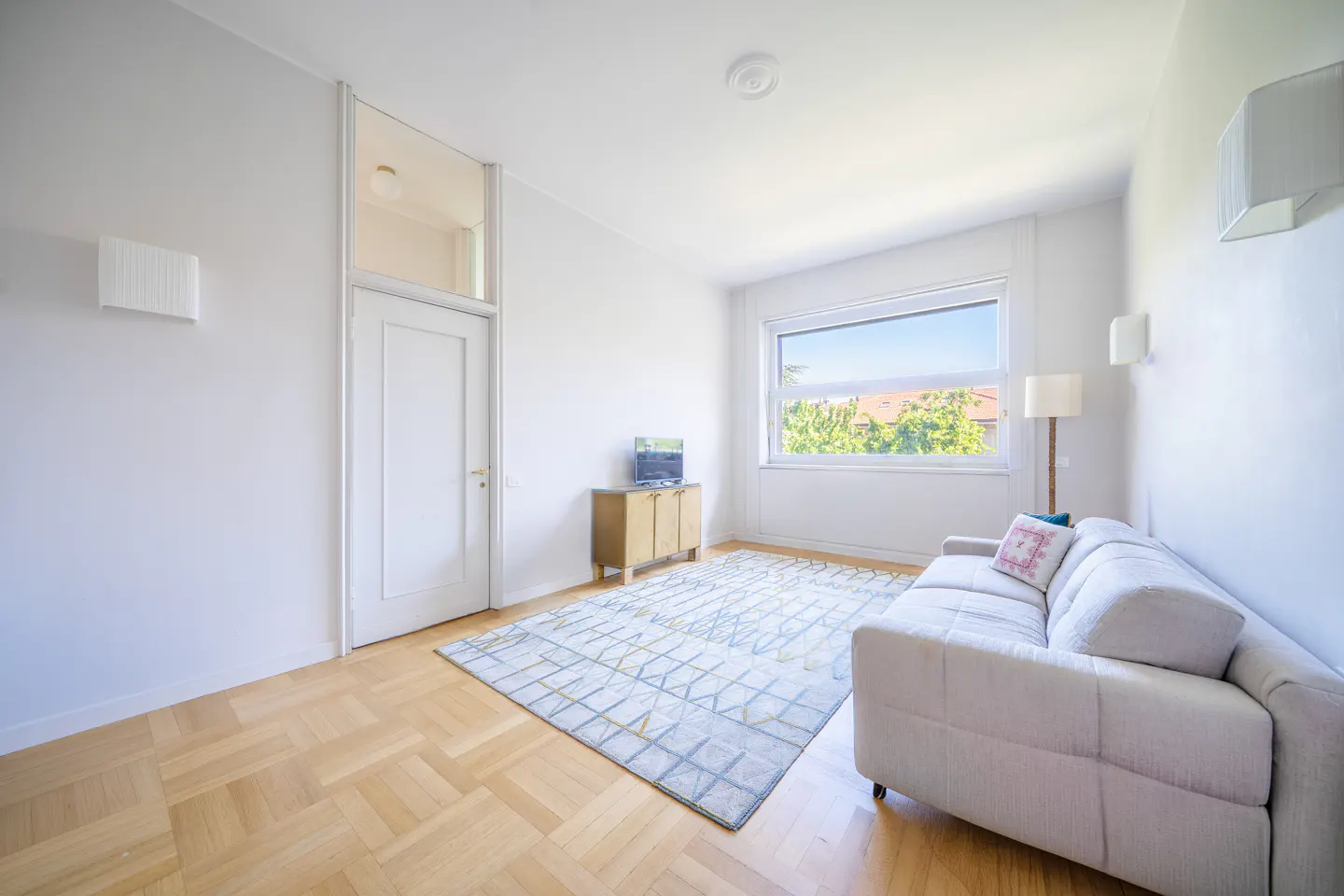 Bright living room with white walls, wood floors, and a large window. A beige sofa sits on a patterned rug, with a floor lamp nearby. A small TV sits on a cabinet.