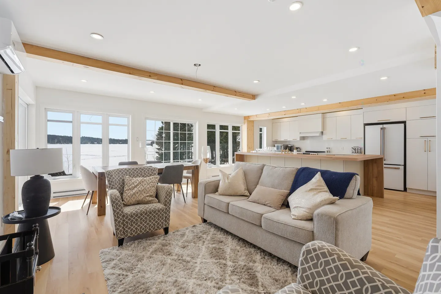 Bright, open-concept living space with a gray sofa, patterned armchairs, and a dining table. Large windows offer a view of a snowy landscape.