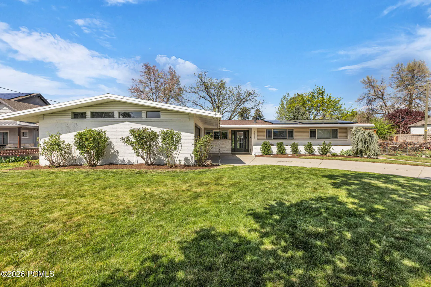 Exterior shot of a single-story white house with a green lawn, blue sky, and scattered trees.