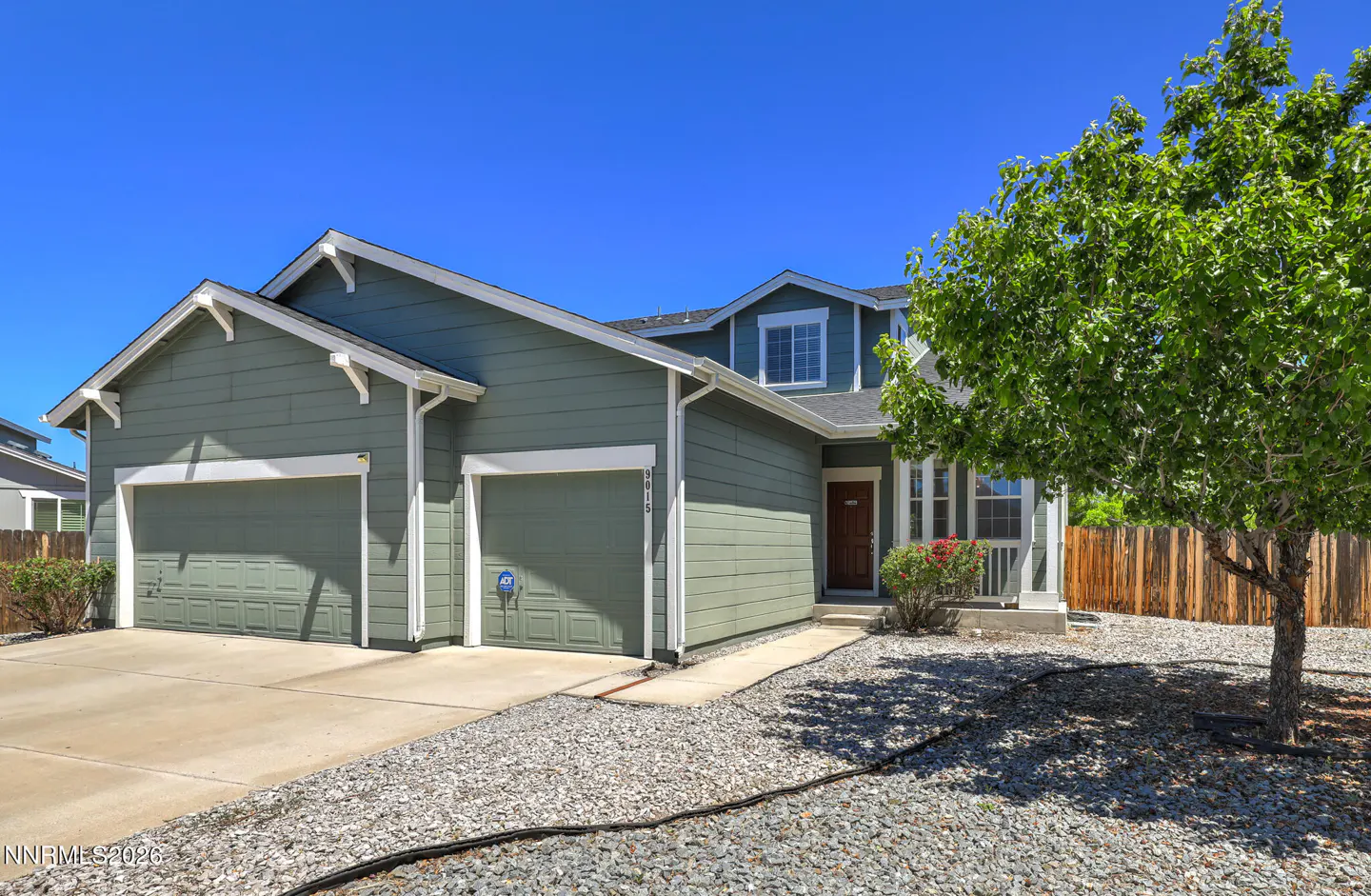 Sage green single-story house with a two-car garage, gravel front yard, and a large tree under a clear blue sky.