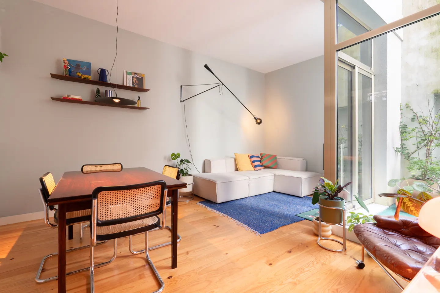 A bright living room with a dining table, white sofa, blue rug, and a large window with plants.