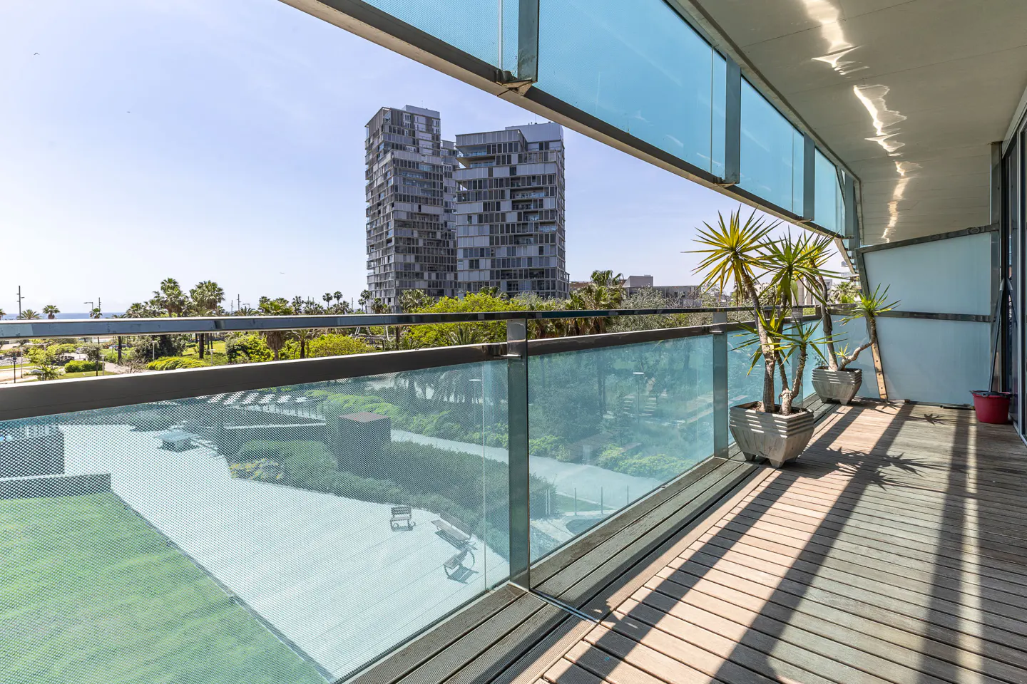 Balcony view with potted plants, wood floor, and glass railings. In the background are tall buildings, trees, and a blue sky.