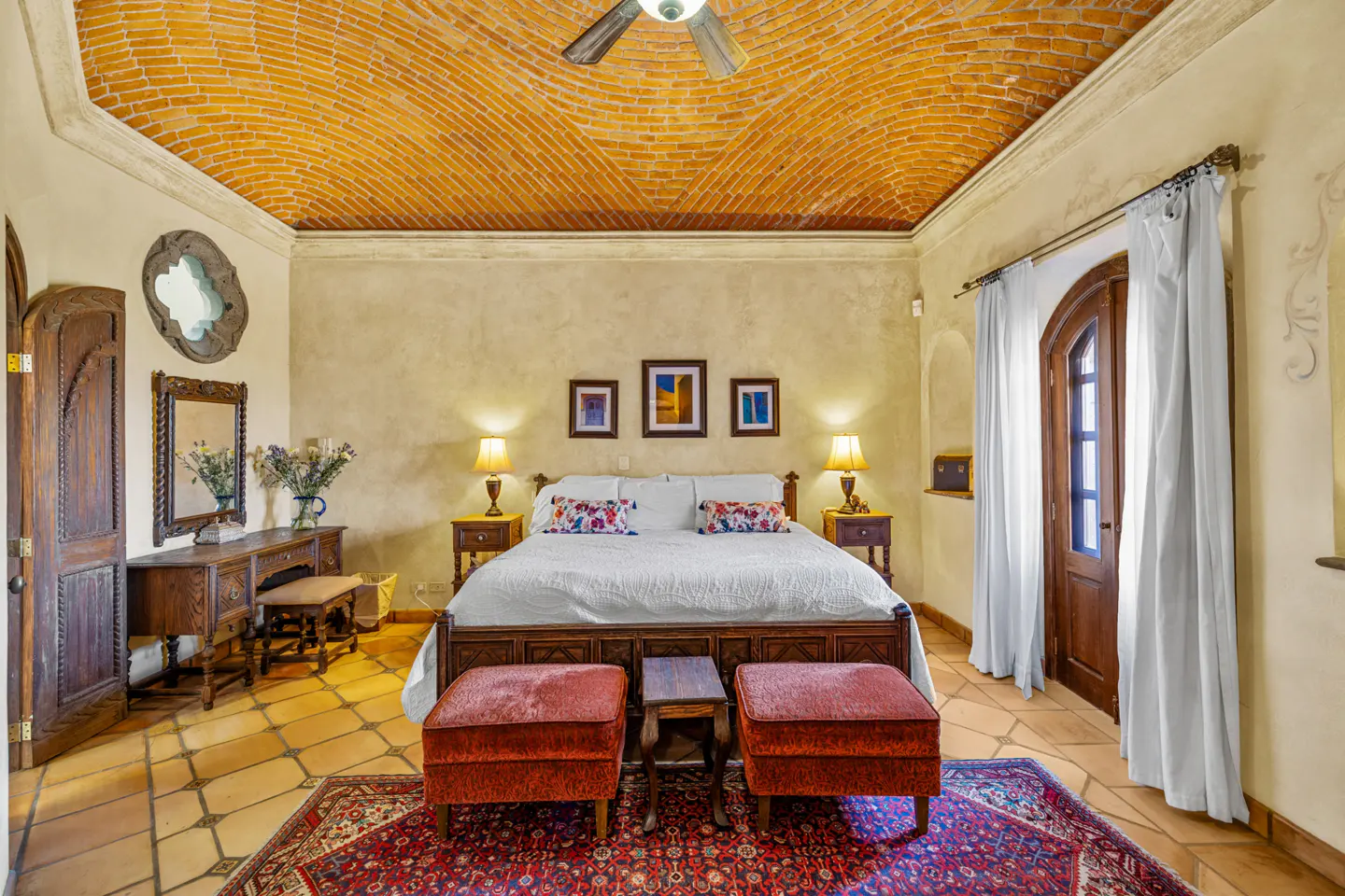 Bedroom with brick ceiling, terracotta tile floor, and a large bed with white linens. Two red ottomans sit at the foot of the bed. A wooden door leads to a balcony.