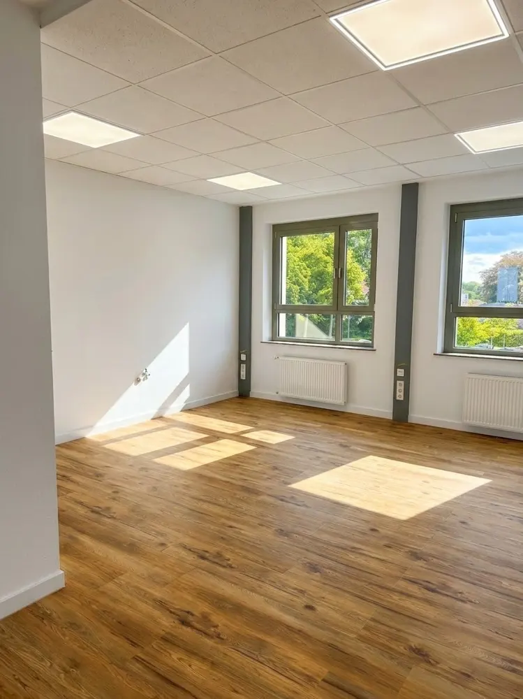 Empty office space with light wood floors, white walls, recessed ceiling lights, and two windows overlooking greenery.
