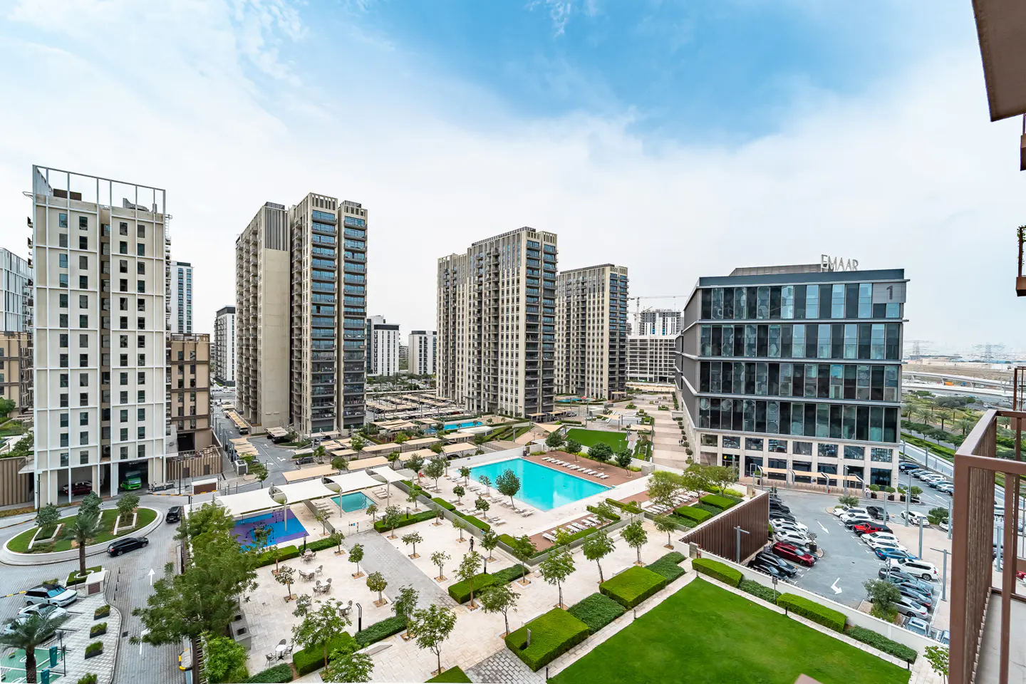 Wide shot of a modern apartment complex with a pool, green spaces, and parking under a cloudy sky.