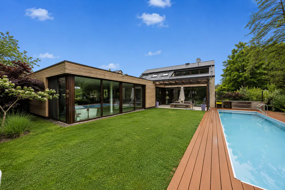 Backyard view of a modern house with a pool, wood deck, green lawn, and glass-walled sunroom under a blue sky.