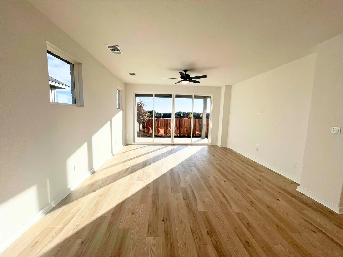 Bright, empty room with light wood floors, white walls, and a black ceiling fan. Sliding glass doors lead to a fenced backyard. Sunlight streams in.