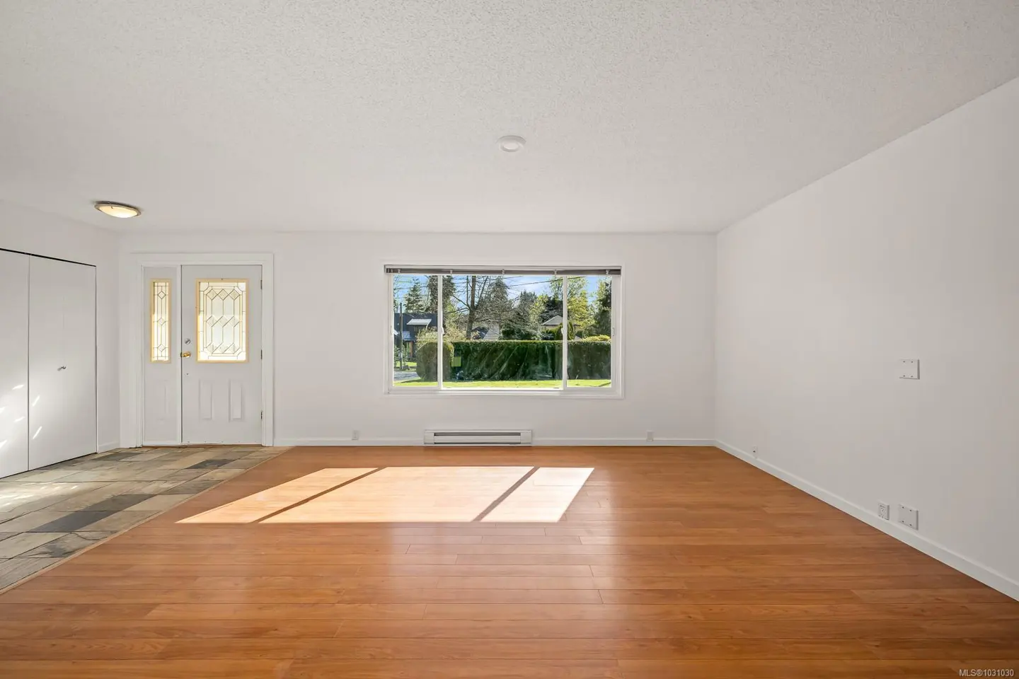 Bright, empty living room with hardwood floors, white walls, and a window overlooking a green yard. A white door with glass panels is on the left.