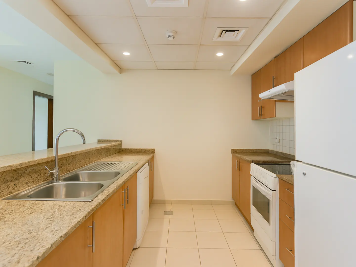 A bright kitchen with beige cabinets, granite countertops, and stainless steel sink. A white refrigerator and oven are on the right.