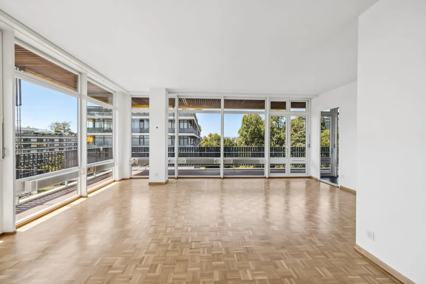 Bright, empty room with parquet floors and white walls. Large windows offer views of balconies, trees, and buildings.