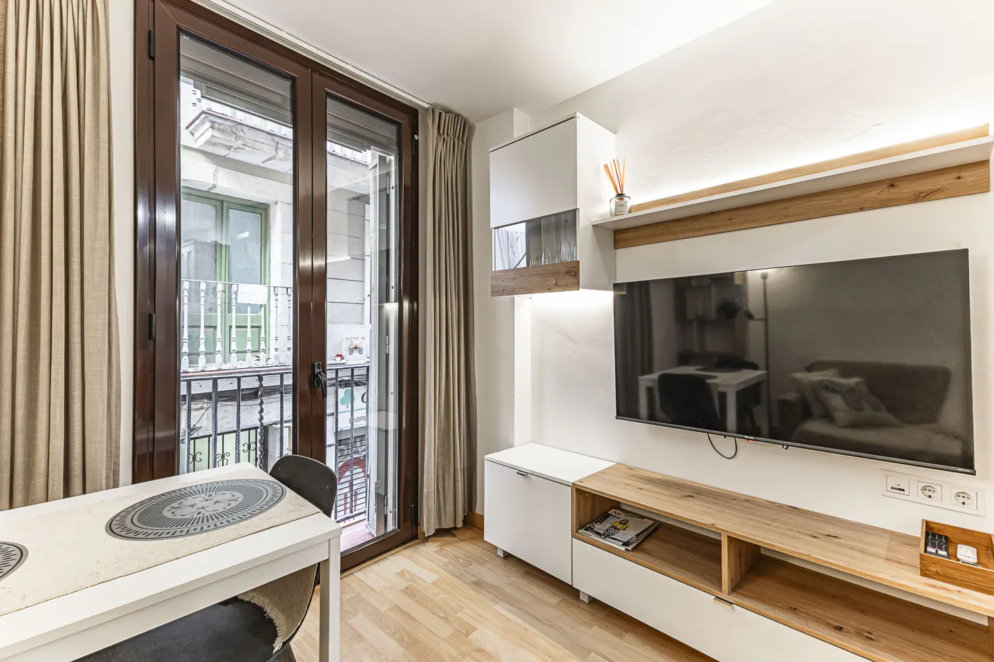 Living room with a white table, chairs, and a large TV on the wall. Balcony doors open to a city view. Neutral color scheme.