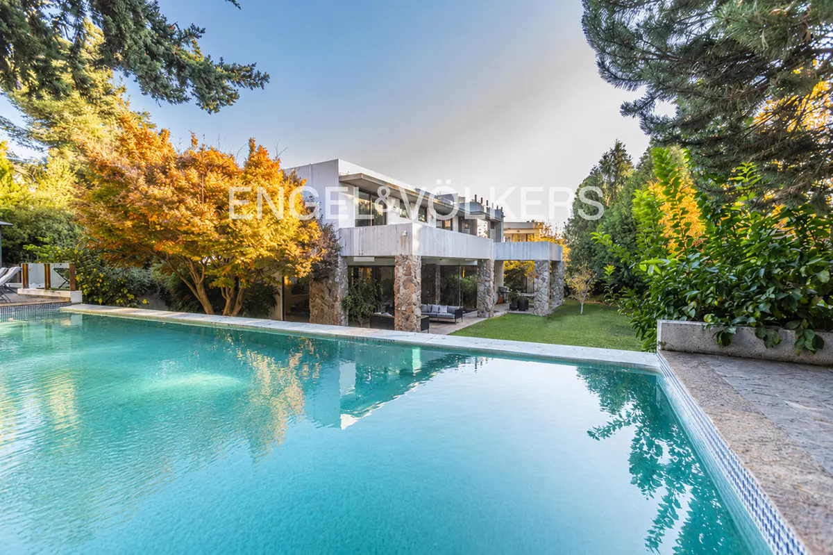 Modern house with a turquoise pool in the foreground. The house has stone pillars and a flat roof, surrounded by trees.