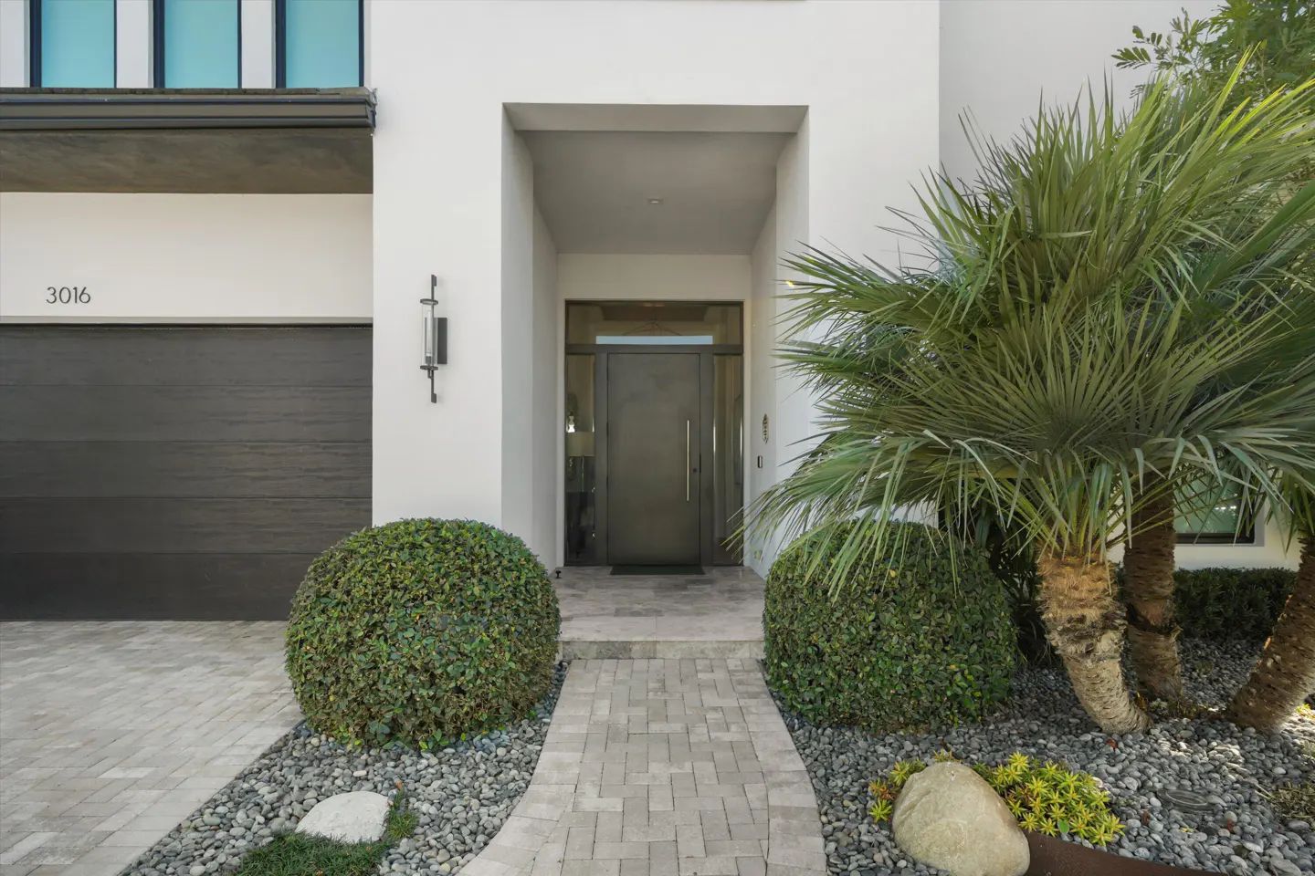 Exterior of a modern home with a gray front door, palm tree, and brick walkway.
