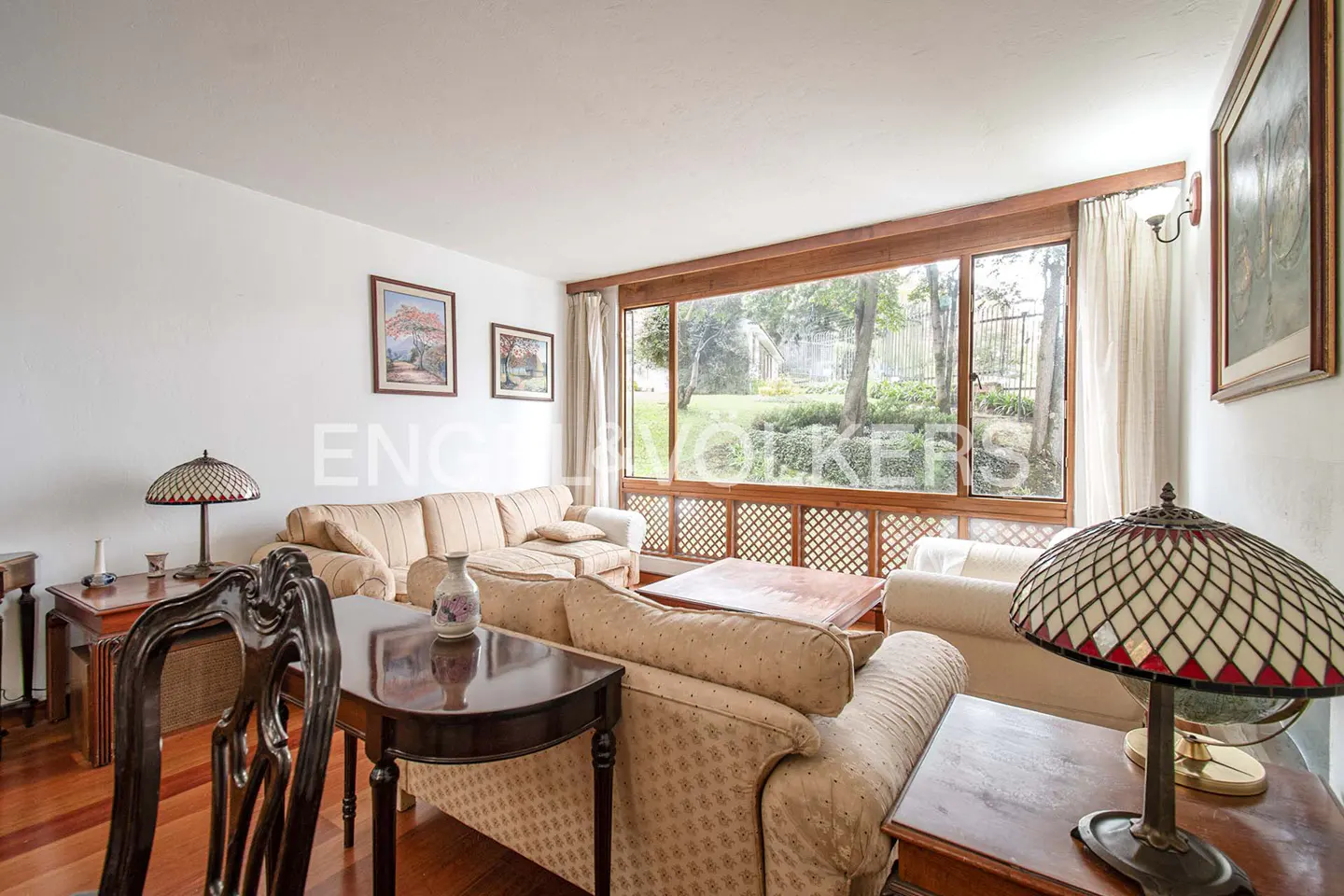 Bright living room with beige sofas, wooden coffee table, stained-glass lamp, and large windows overlooking a garden.