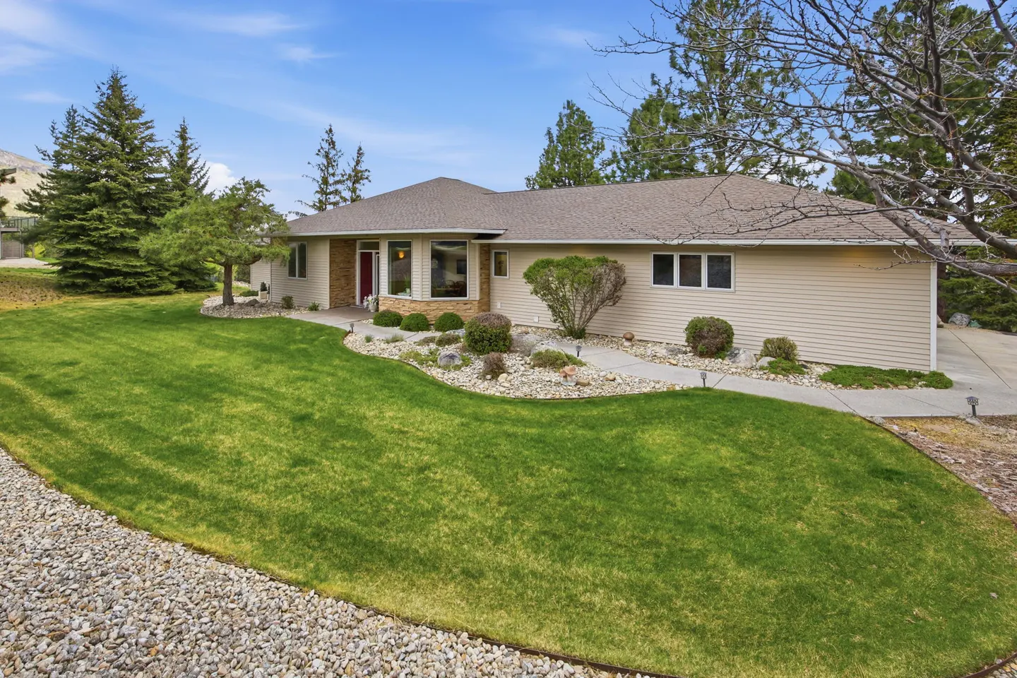 A tan ranch-style house with a red door, green lawn, and rock landscaping on a sunny day.