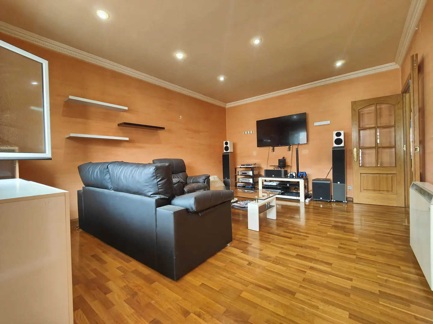 Living room with wood floors, orange walls, and recessed lighting. A black leather sofa faces a TV and speakers. Shelves are on the left wall.