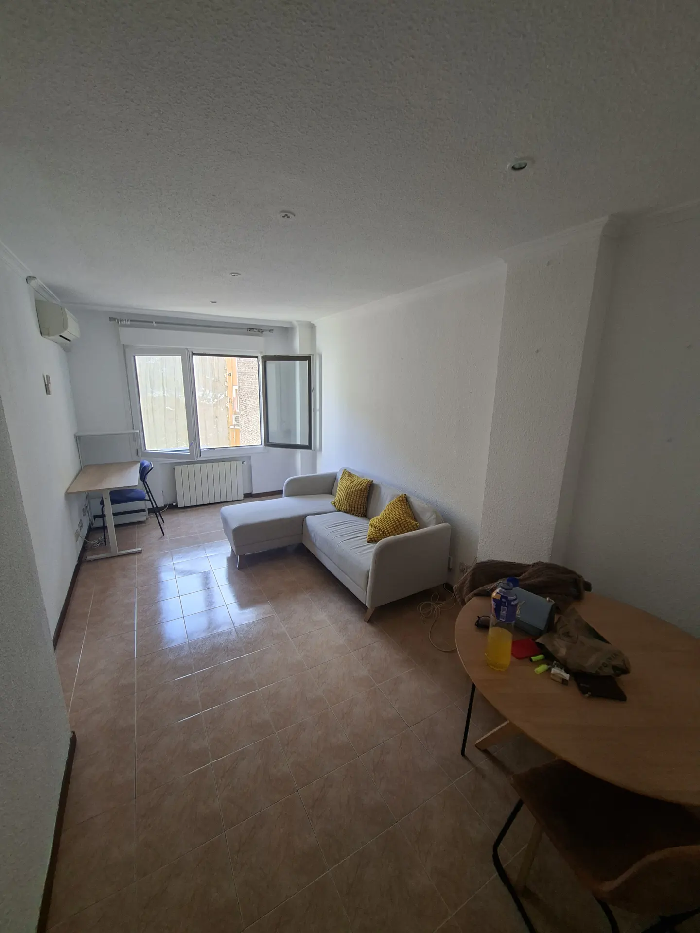 Bright living room with tile floor, white walls, and a gray sectional sofa with yellow pillows. A desk sits near a window, and a round table is in the foreground.