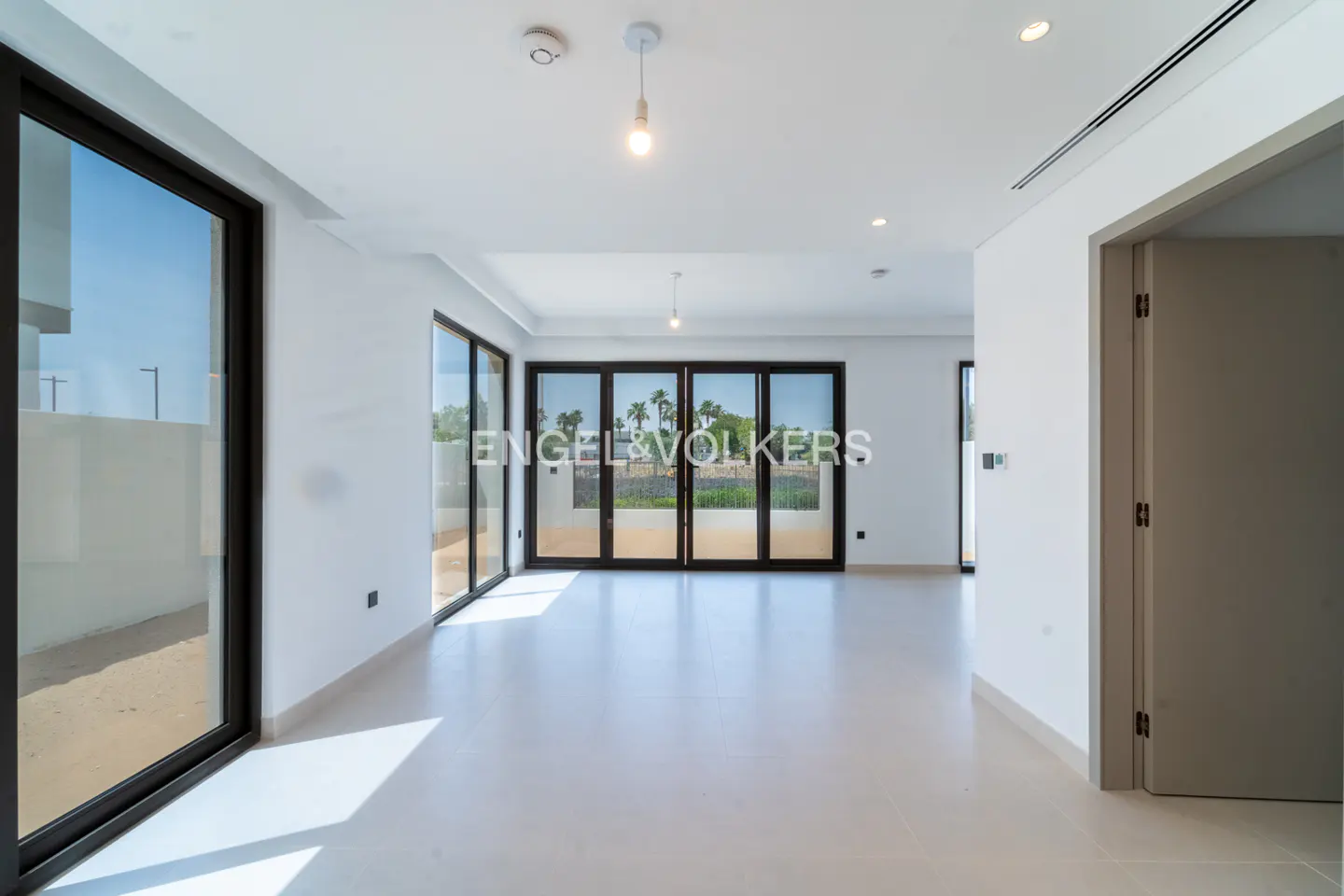 Bright, empty room with white walls and floors. Large black-framed windows offer outdoor views. A doorway is visible on the right.