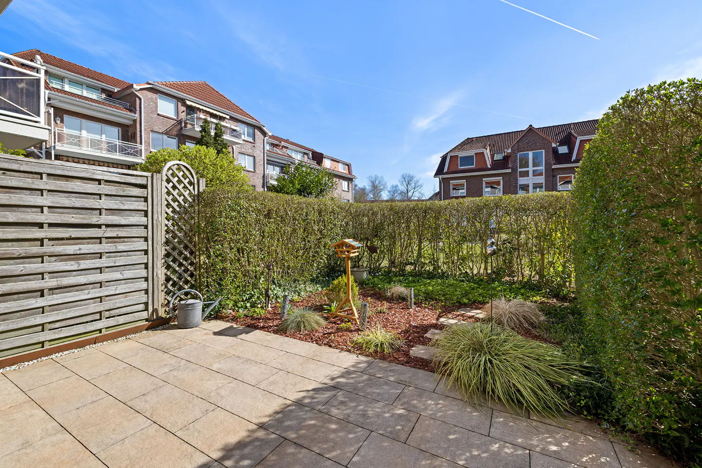 A patio with a wooden fence, a watering can, and a garden with a birdhouse, shrubs, and mulch. Buildings are visible in the background.