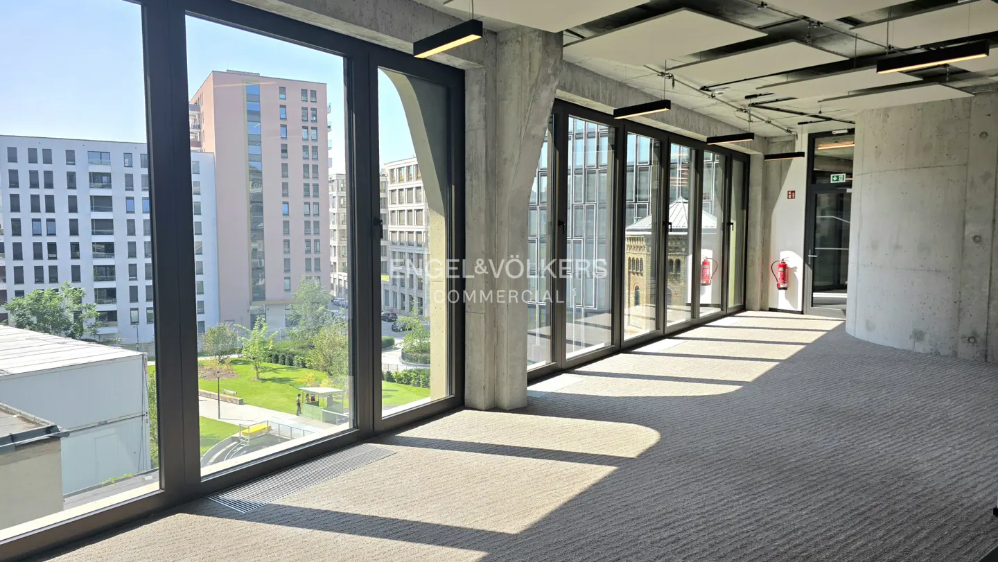 Modern office hallway with floor-to-ceiling windows, concrete walls, carpeted floor, and a view of city buildings.
