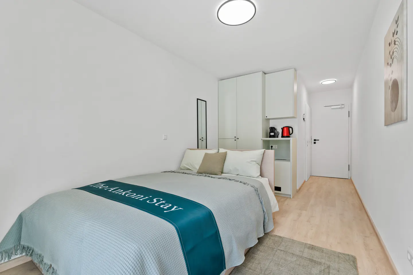 Bright studio apartment with a bed, white cabinets, and a small kitchenette. A teal runner with white text is on the bed.
