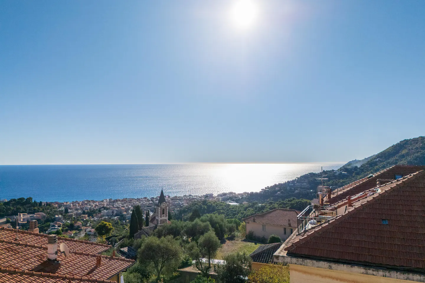 Scenic view of a coastal town with a church steeple, red-tiled roofs, and the blue sea under a bright, sunny sky.