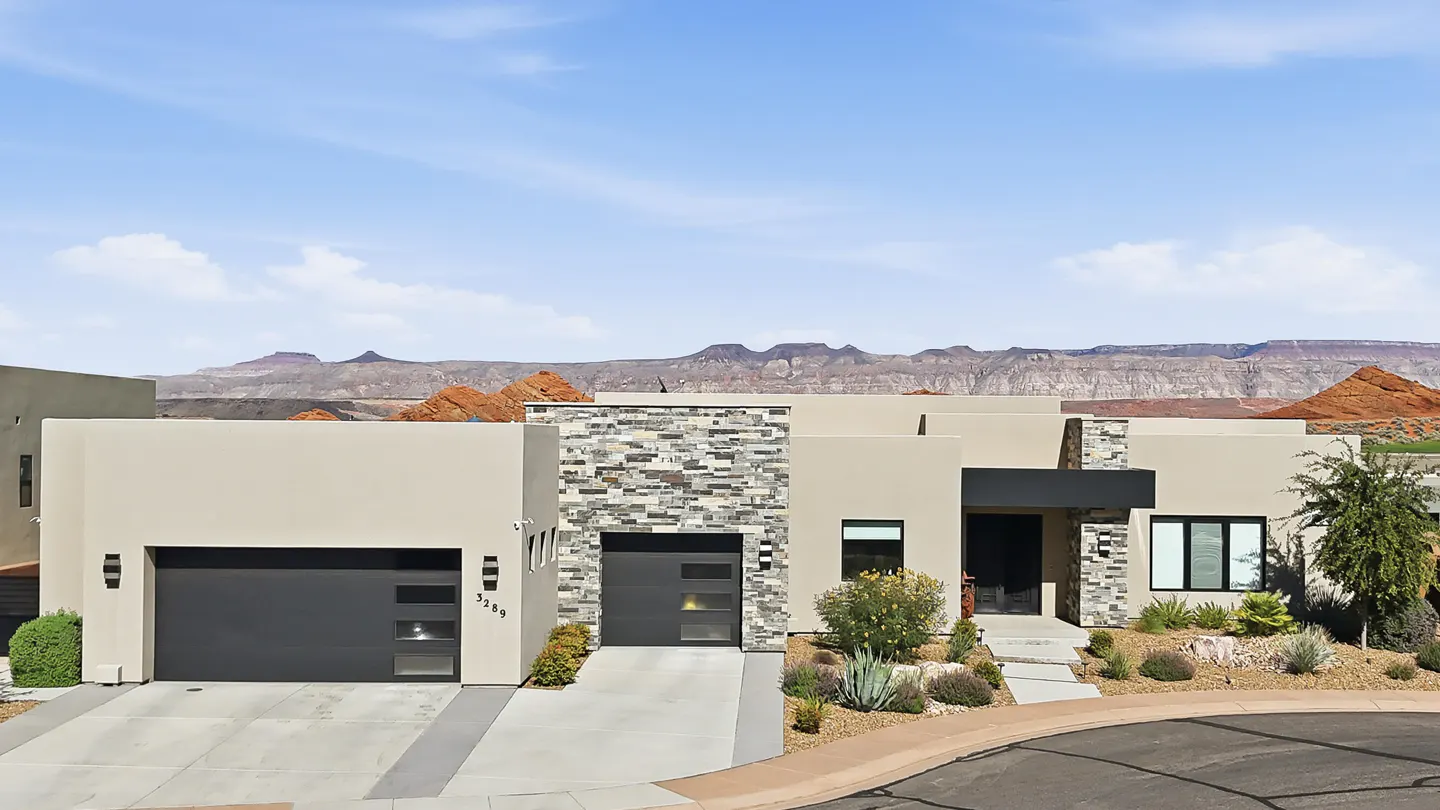 Modern desert home with tan stucco, stone accents, two garages, and desert landscaping against a mountain backdrop.