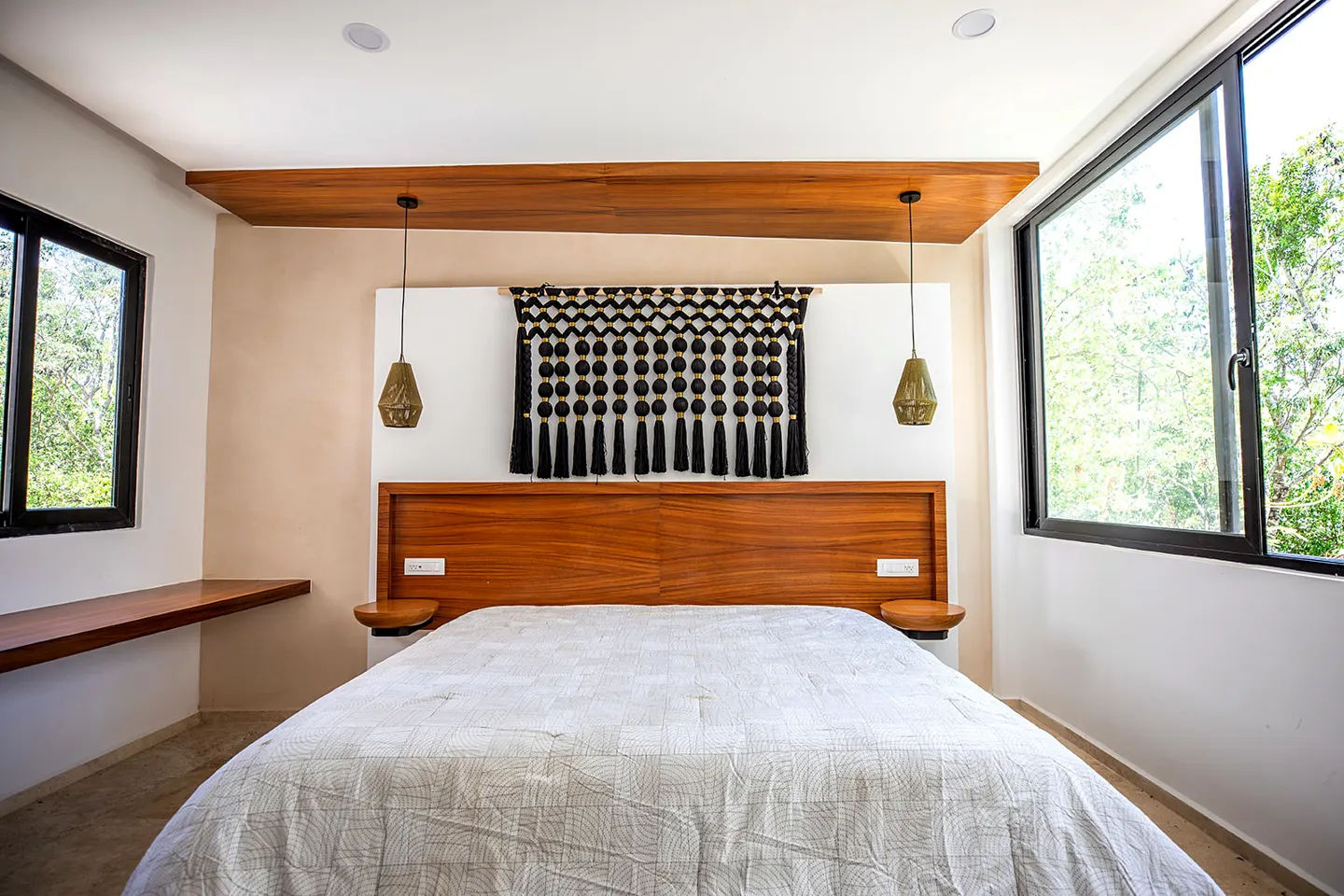 Bedroom with a wooden headboard, white patterned quilt, and black tassel wall hanging. Large windows let in natural light.